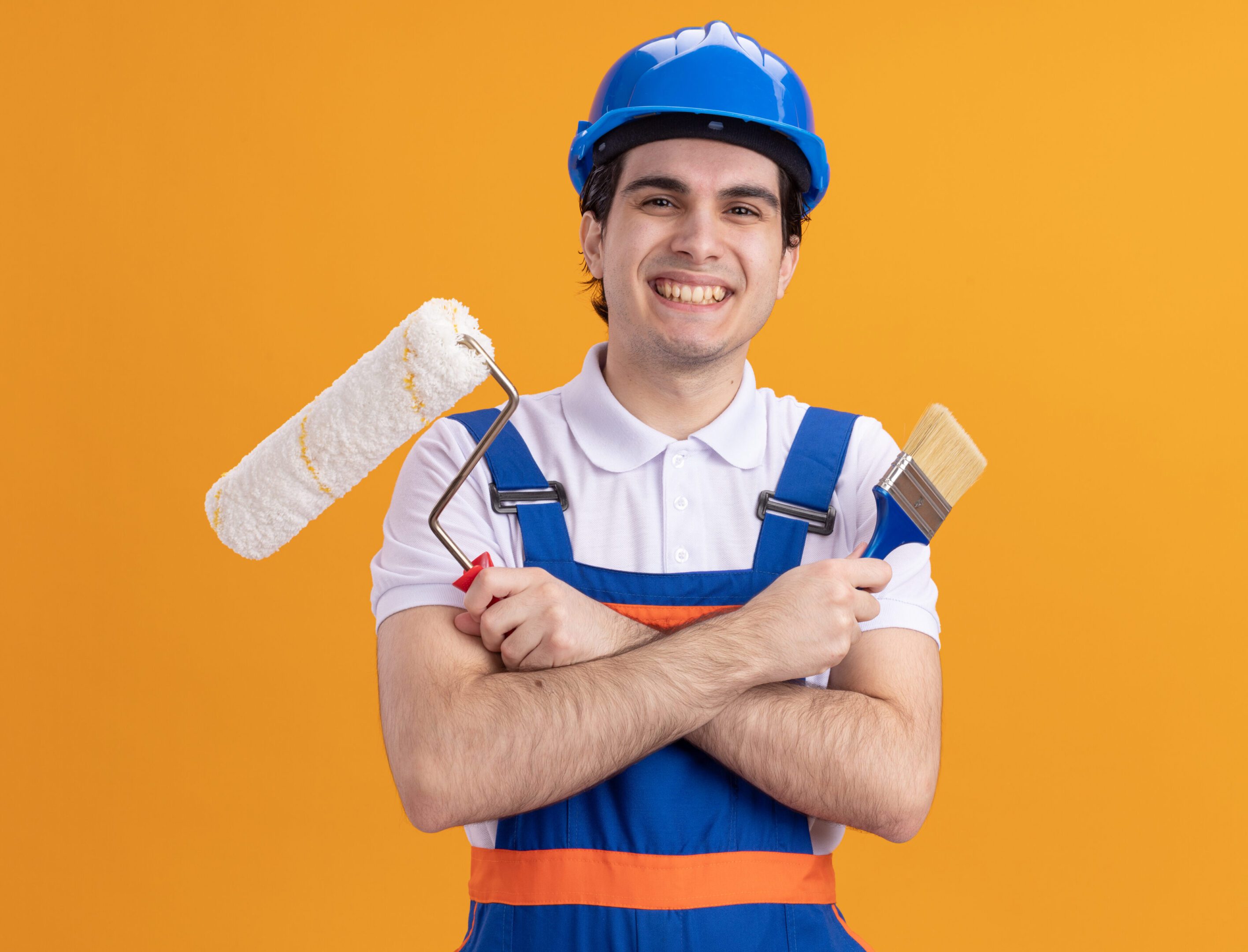 young builder man in construction uniform and safety helmet holding paint brush and roller looking at camera with big smile on face standing over orange background young builder man in construction uniform and safety helmet holding paint brush and roller looking at camera with big smile on face standing over orange background